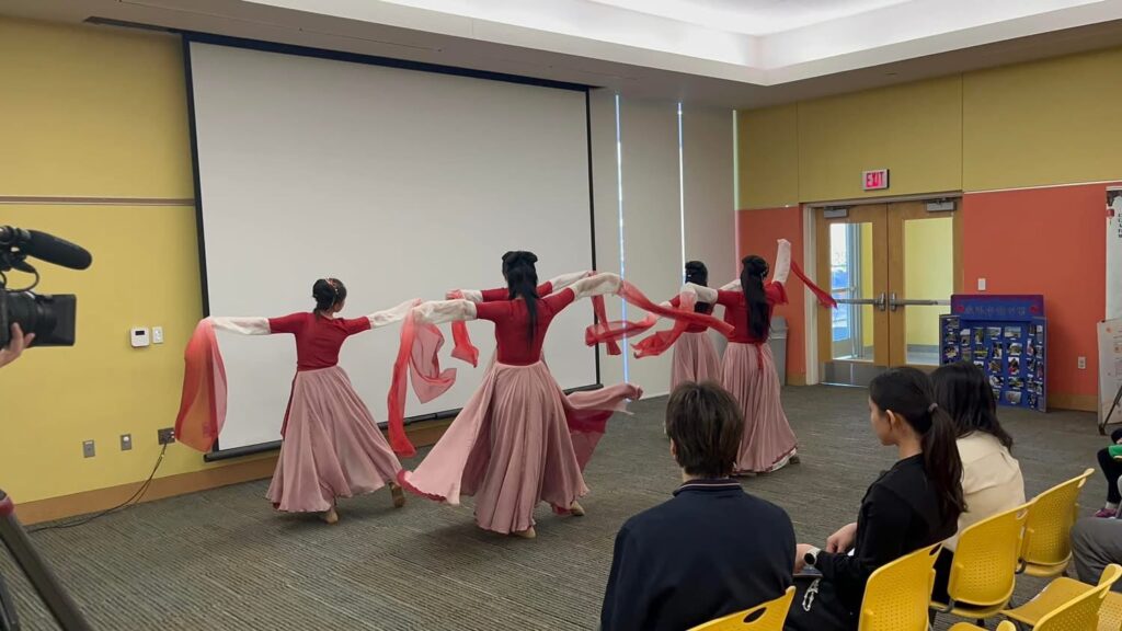 Four dancers in flowing pink skirts and red tops perform a ribbon dance indoors, facing away from the audience. They hold long pink-and-white ribbons, while audience members watch from yellow chairs.