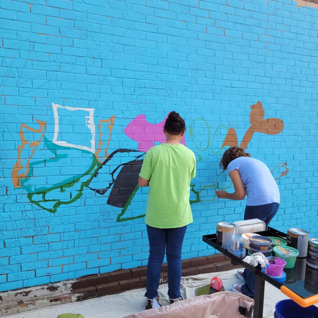 Two people painting a mural onto a blue wall.