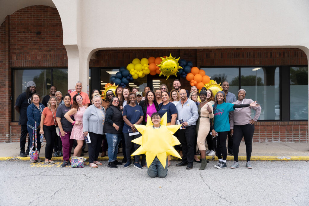 Several dozen people standing in front of a storefront in front of orange, yellow, and black balloons, with a person dressed in a sun costume kneeling in front.
