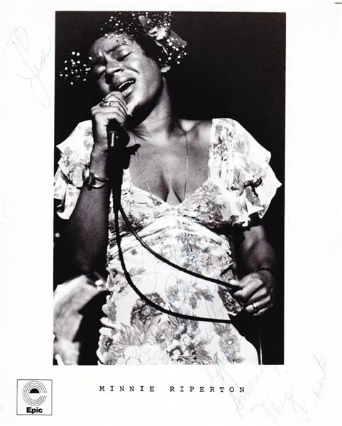Dark-skinned woman closes her eyes and sings into a chorded microphone while wearing a floral dress and flowers in her hair.
