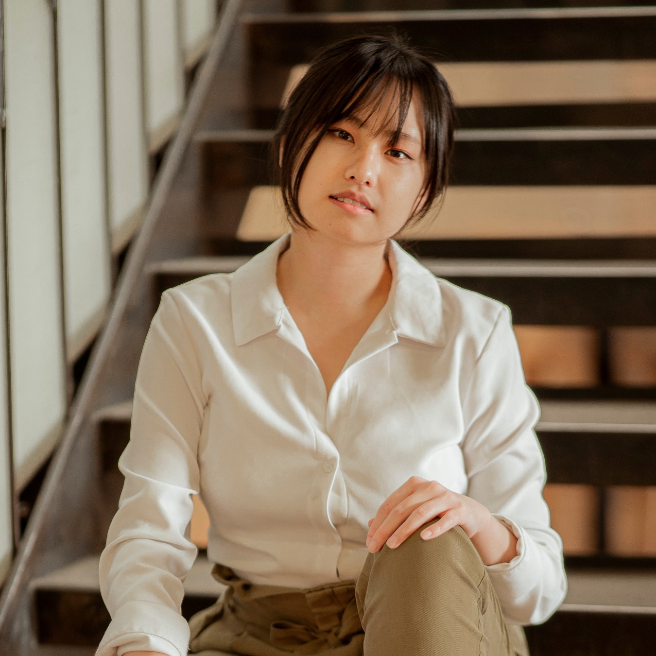 Headshot of a person of medium light skin tone with black hair tied back, wearing a white button-down shirt and tan pants.