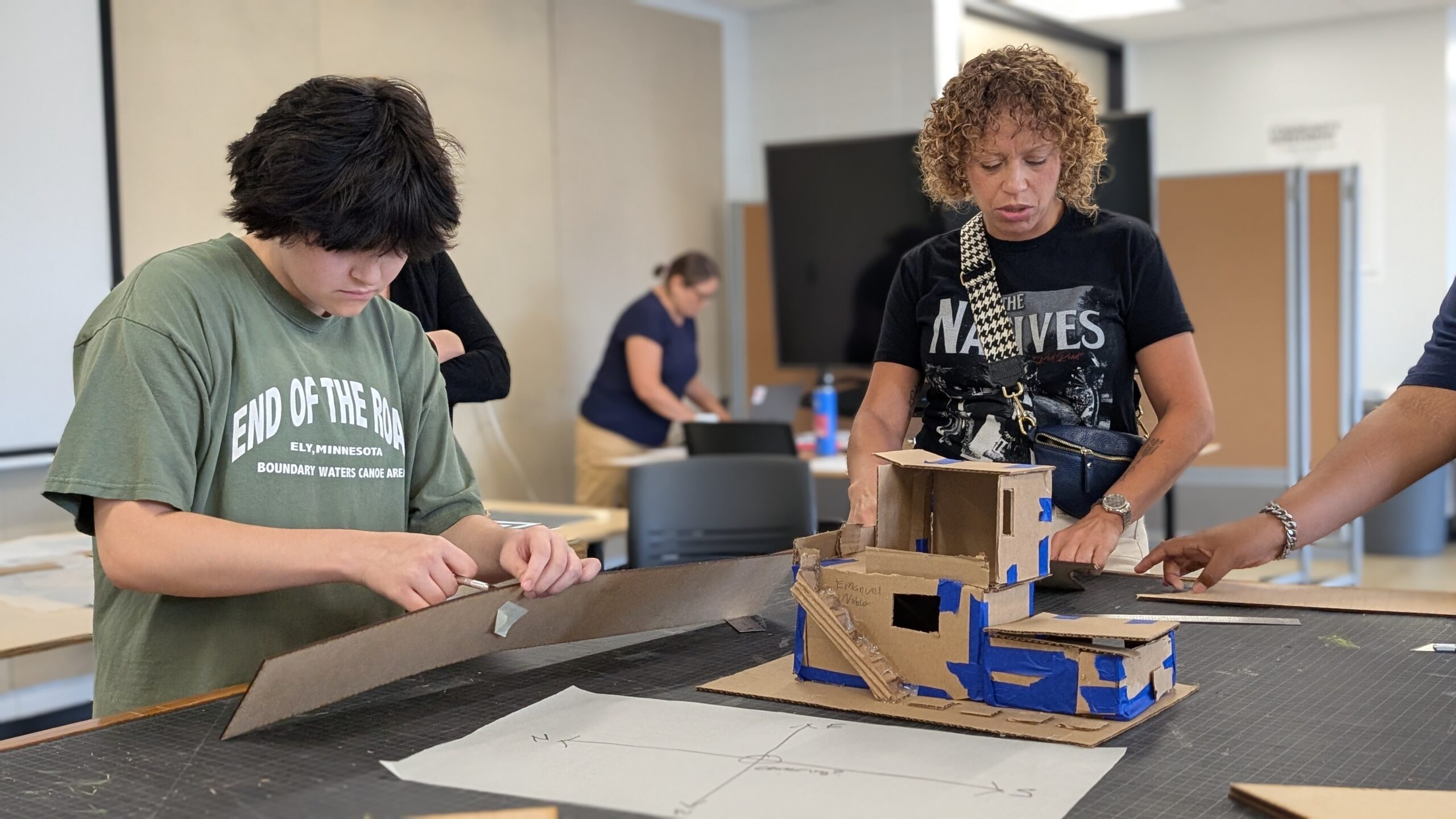 An adult and a younger person standing at a large table while working on cardboard models of building structures.