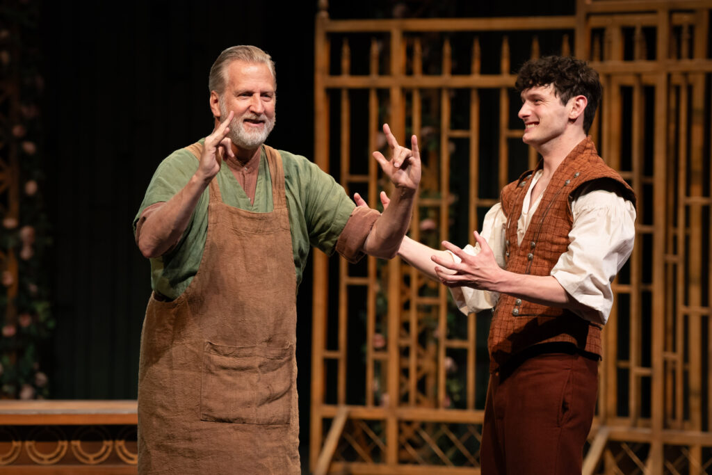 Two light-skinned people sign with their hands while standing in front of a wooden framed set.