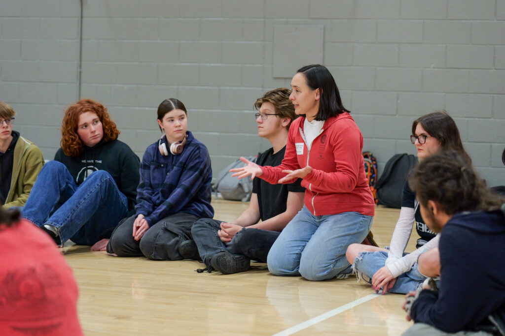 woman in a red hoodie speaks animatedly while seated on a gym floor with a circle of attentive high school students.