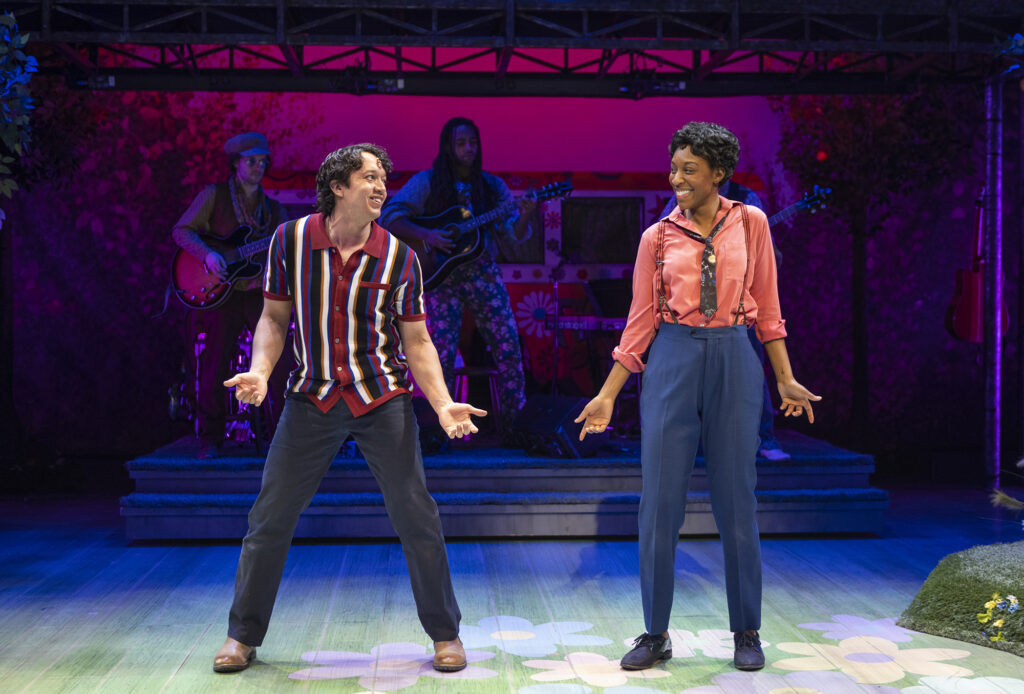 Two people perform on stage, with their arms extended out from their hips and smiling at each other. In the background, two people wearing sunglasses play guitar on a stage.