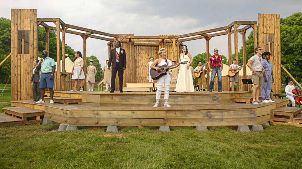 Multiple people perform on a wooden stage on a lawn outside, some singing and some playing guitars.