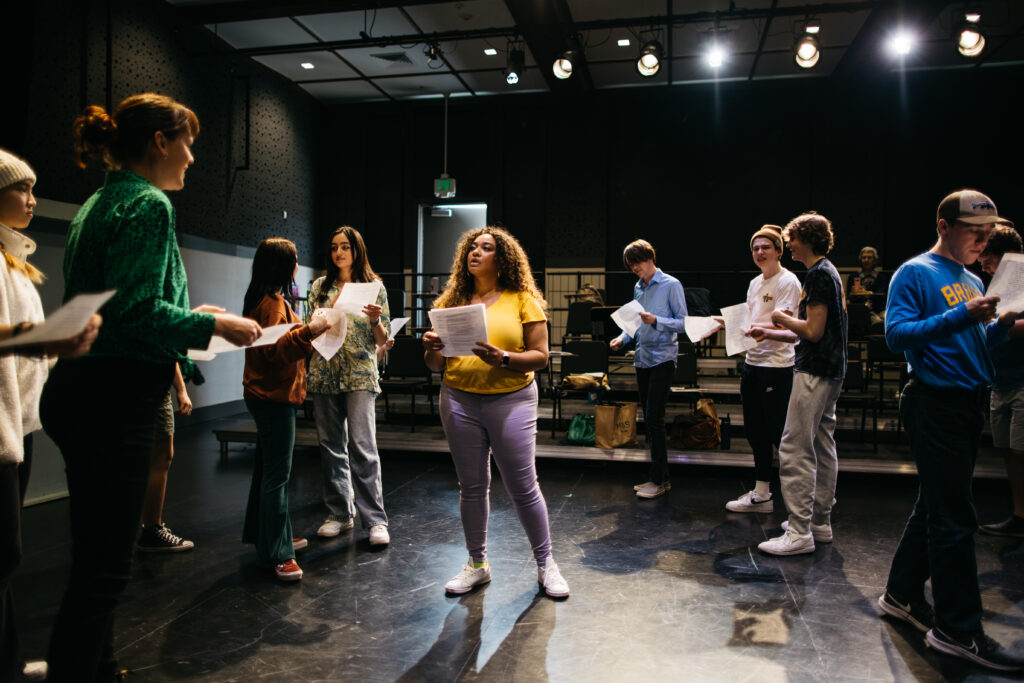 A group of high school students hold script papers in a black box theater.