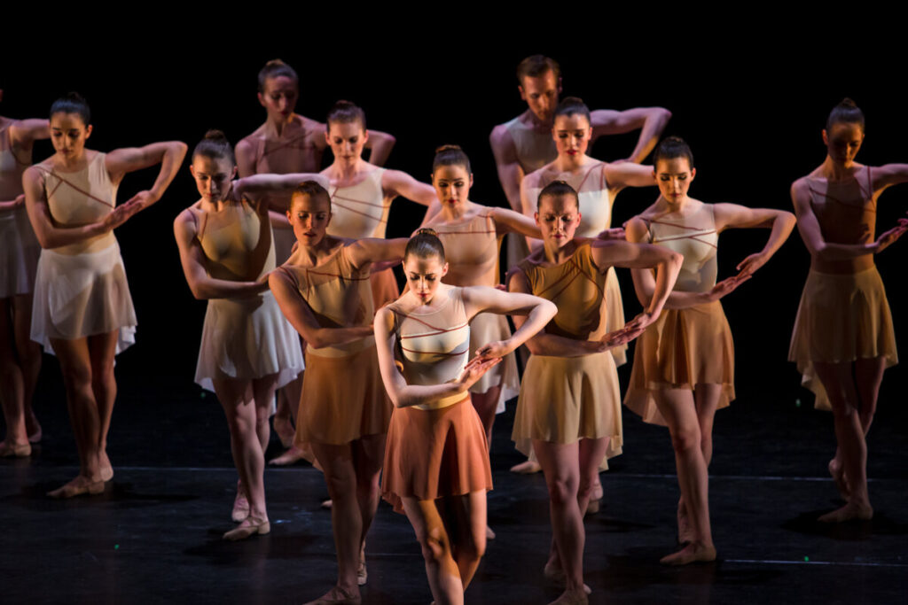 A group of dancers in white and brown costumes cradling their arms and looking downwards.