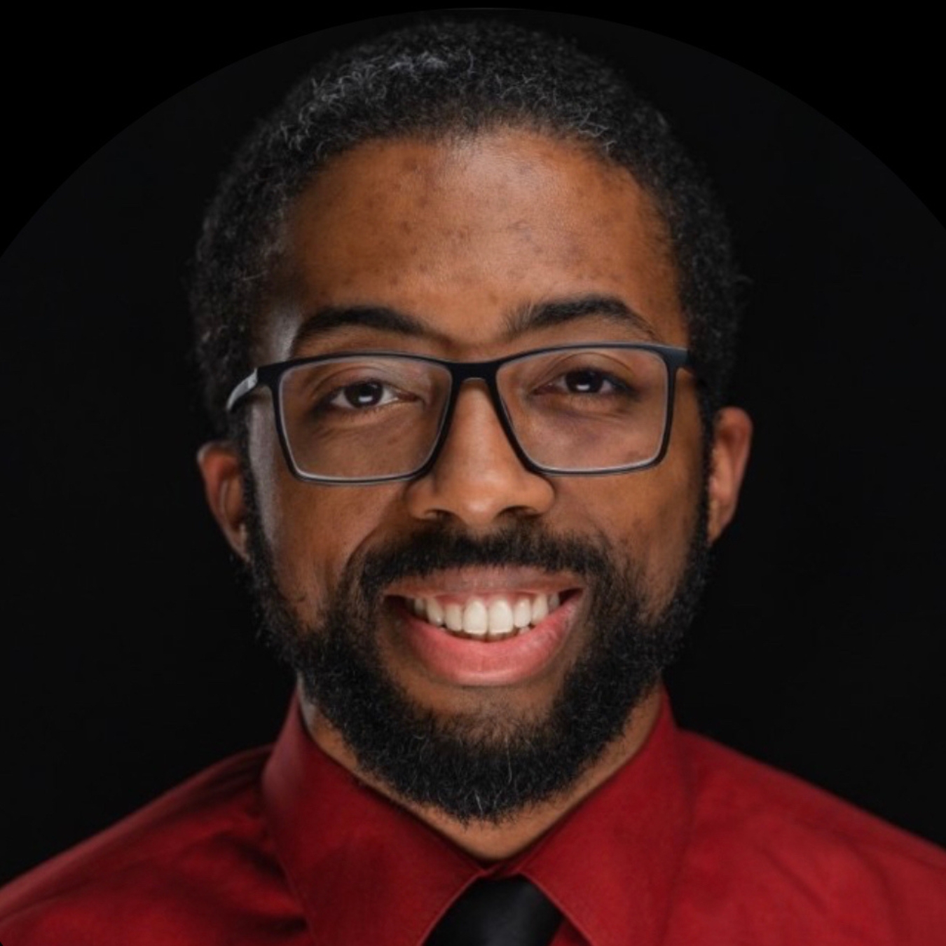 Headshot of a person of dark skin tone with black hair, glasses, and a beard, wearing a red button-down shirt and black tie.