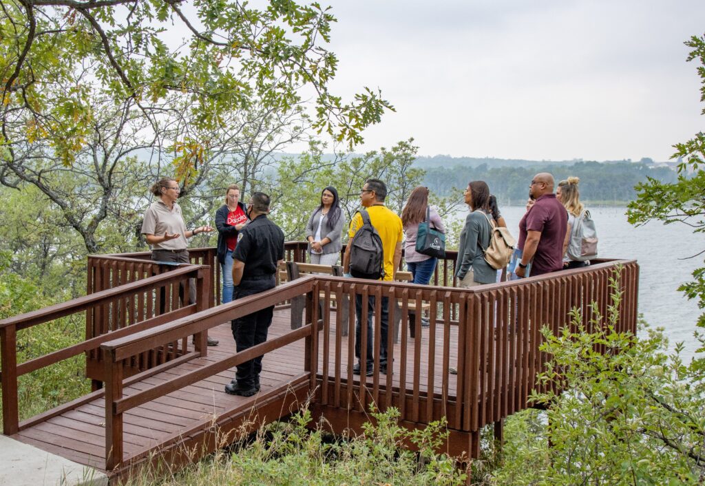A group of people stand on an overlook and listen to a person talking and gesturing. The background of the image is a river with trees at a distance.