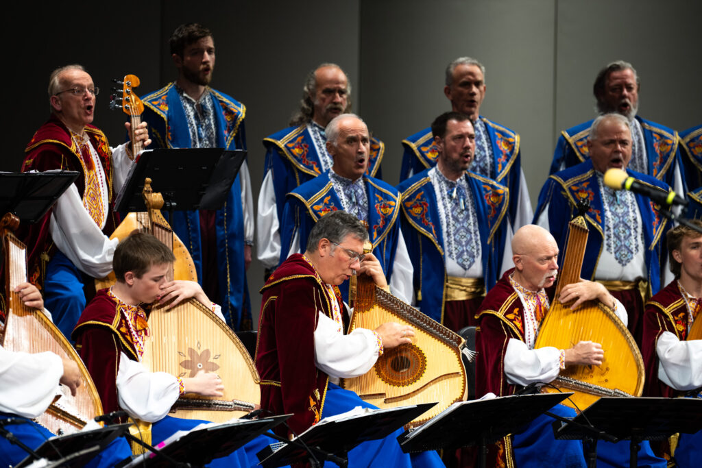 Men in traditional wear sit and stand on a stage with stringed instruments in the front row.