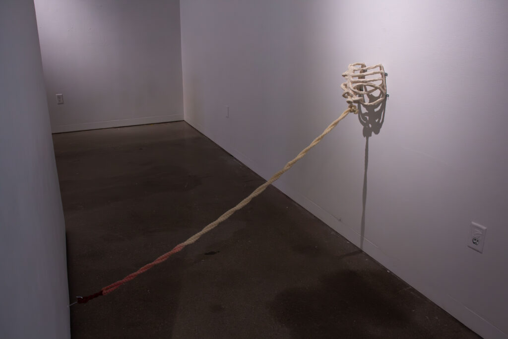 A ceramic ribcage attached to a gallery wall with yarn leading from the breastbone to the floor.