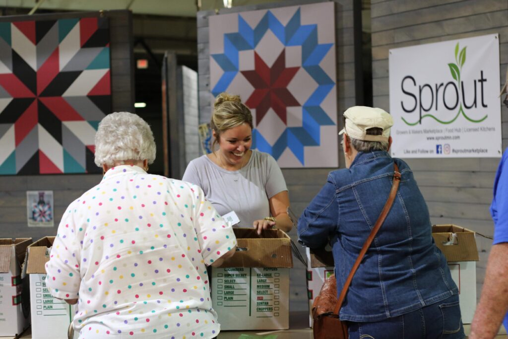A smiling person stands behind a table with open cardboard boxes on it, facing two people who have their backs to the camera and are reaching into the boxes. In the background there's a banner that reads "Sprout," and two barn quilt designs hanging on the wall.
