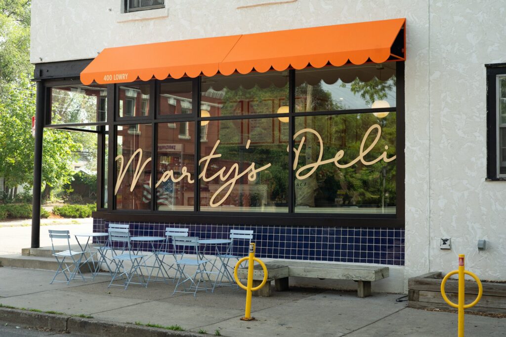A corner business with white siding and an orange awning reads, in gold cursive lettering, "Marty's Deli" on a window.