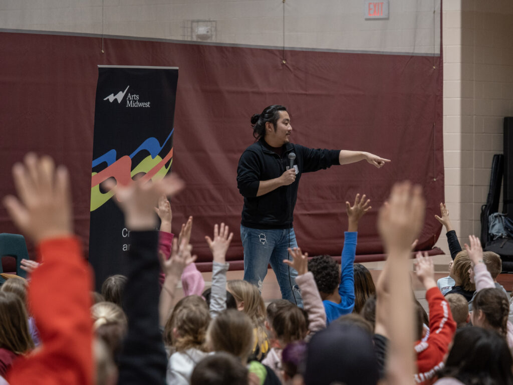 A man holding a microphone points at a crowd of students seated with their arms raised.