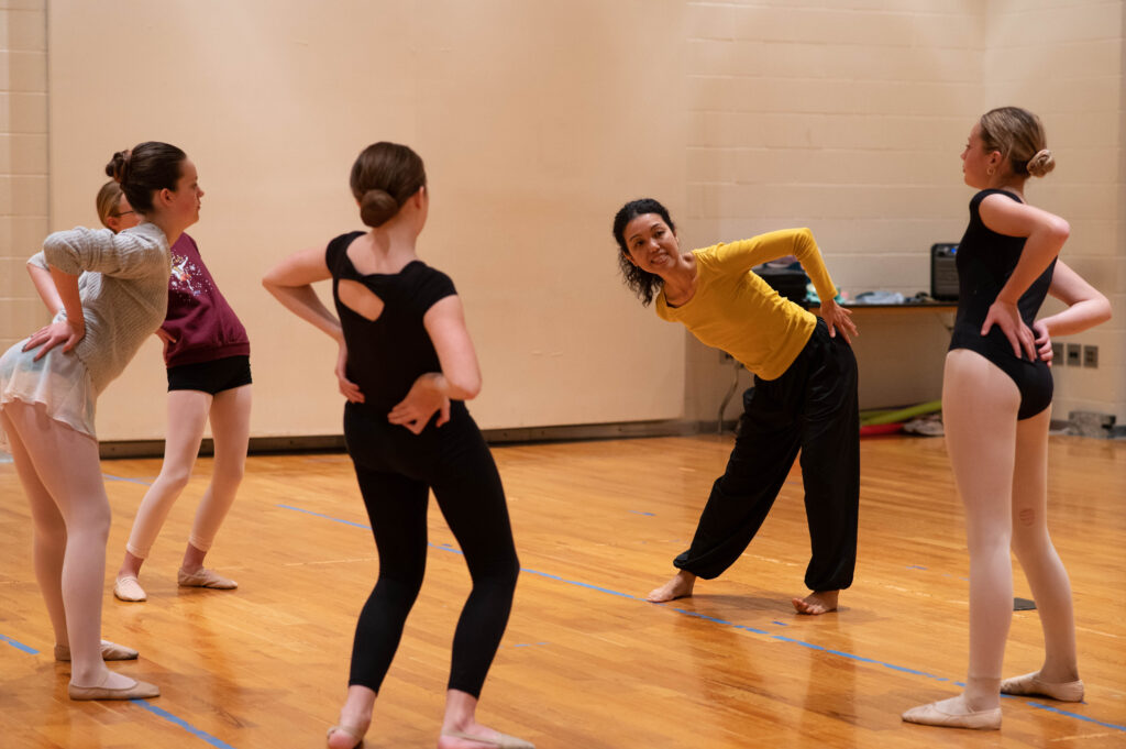 A dance instructor in a yellow shirt leans to the side with hands on hips, smiling at a small group of ballet students who mirror her pose in a studio with a wood floor.