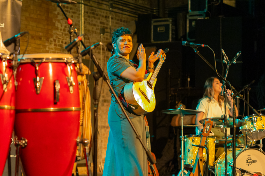 A musician on stage smiles and claps while holding a small stringed instrument, framed by microphones and vibrant lighting. A drummer performs beside her.