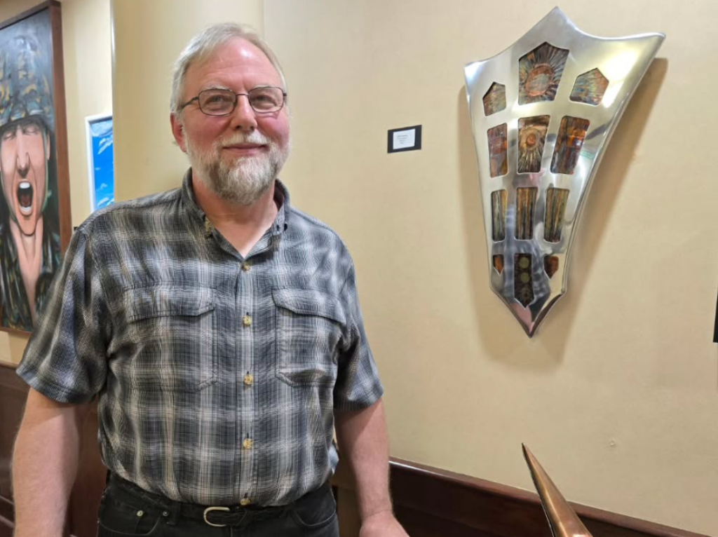 Light-skinned man in button-down smiles and stands next to artwork in a room.