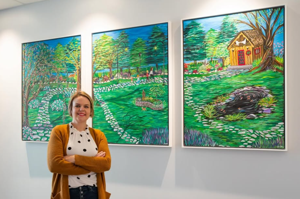 Light-skinned woman crosses her arms and stands in front of three green landscape paintings of a yard and garden.