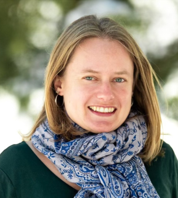 Headshot of a smiling person of light skin tone with medium length blonde hair, wearing a patterned blue scarf over a dark green shirt.