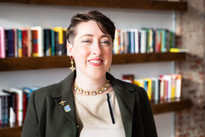 Headshot of a smiling person of light skin tone and short brown hair slicked to the side, wearing earrings and a necklace with gold accents, and a tan shirt under a black blazer with two small pins in the shape of an airplane and a book.