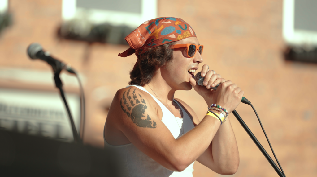 A person wearing a patterned bandana on their head and orange sunglasses singing into a microphone.