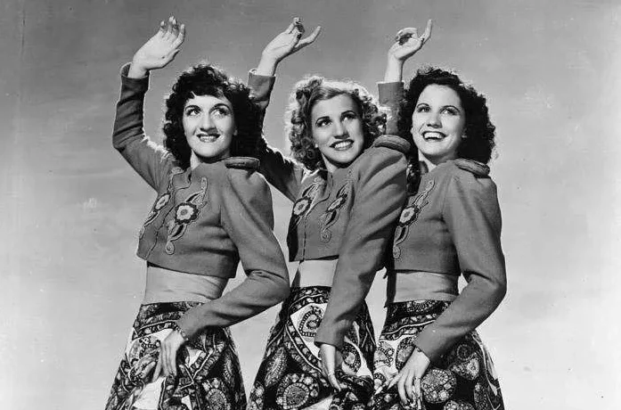 Three light-skinned women smile with their hand up and look to the side.